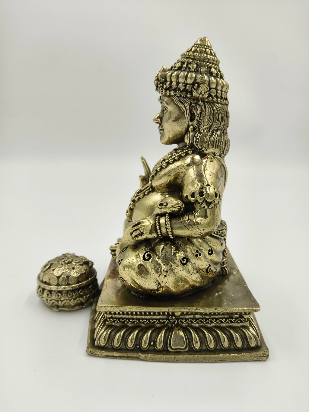 Side view of medium brass Kubera statue seated on decorative base with small coin container