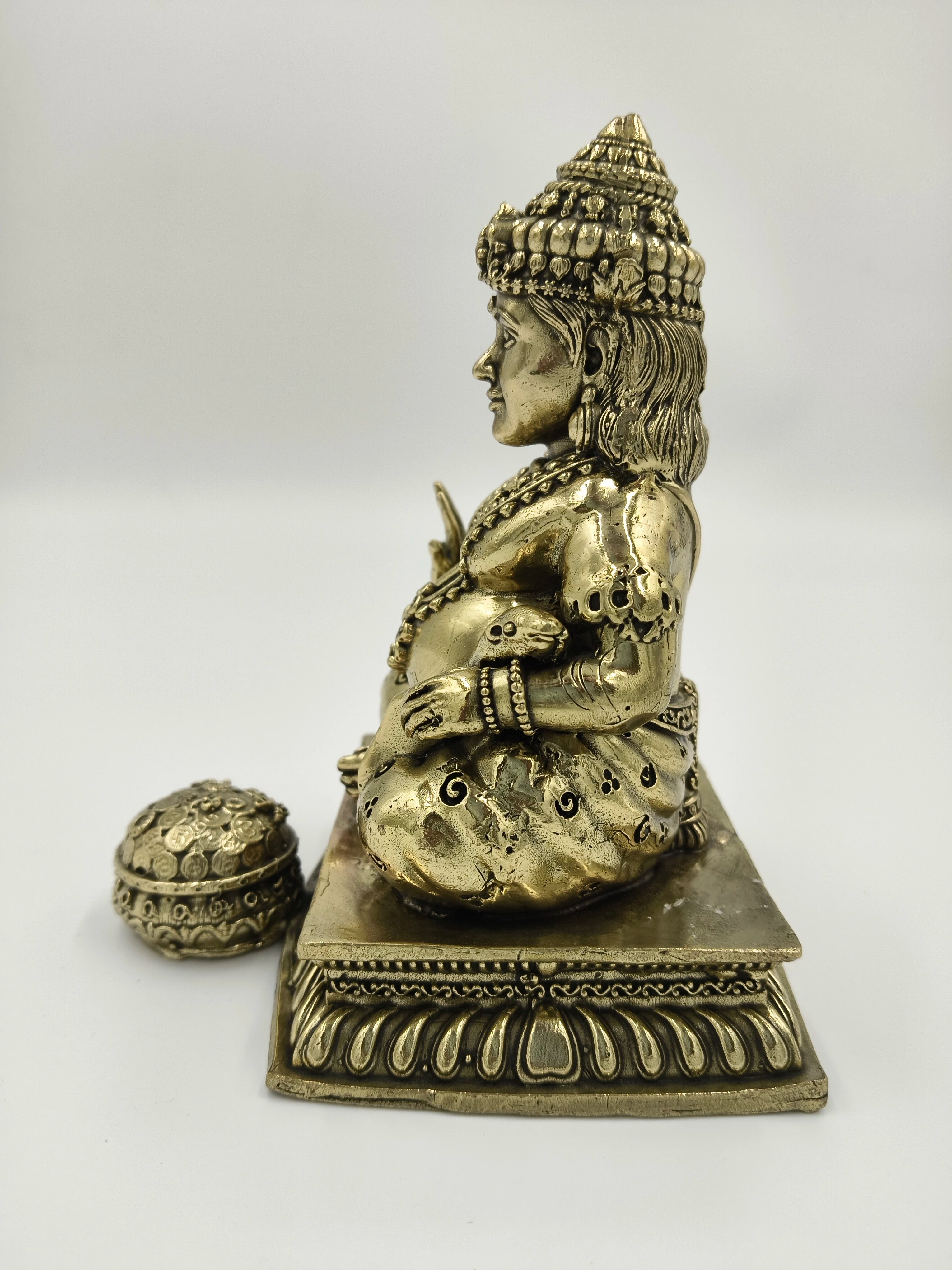 Side view of medium brass Kubera statue seated on decorative base with small coin container