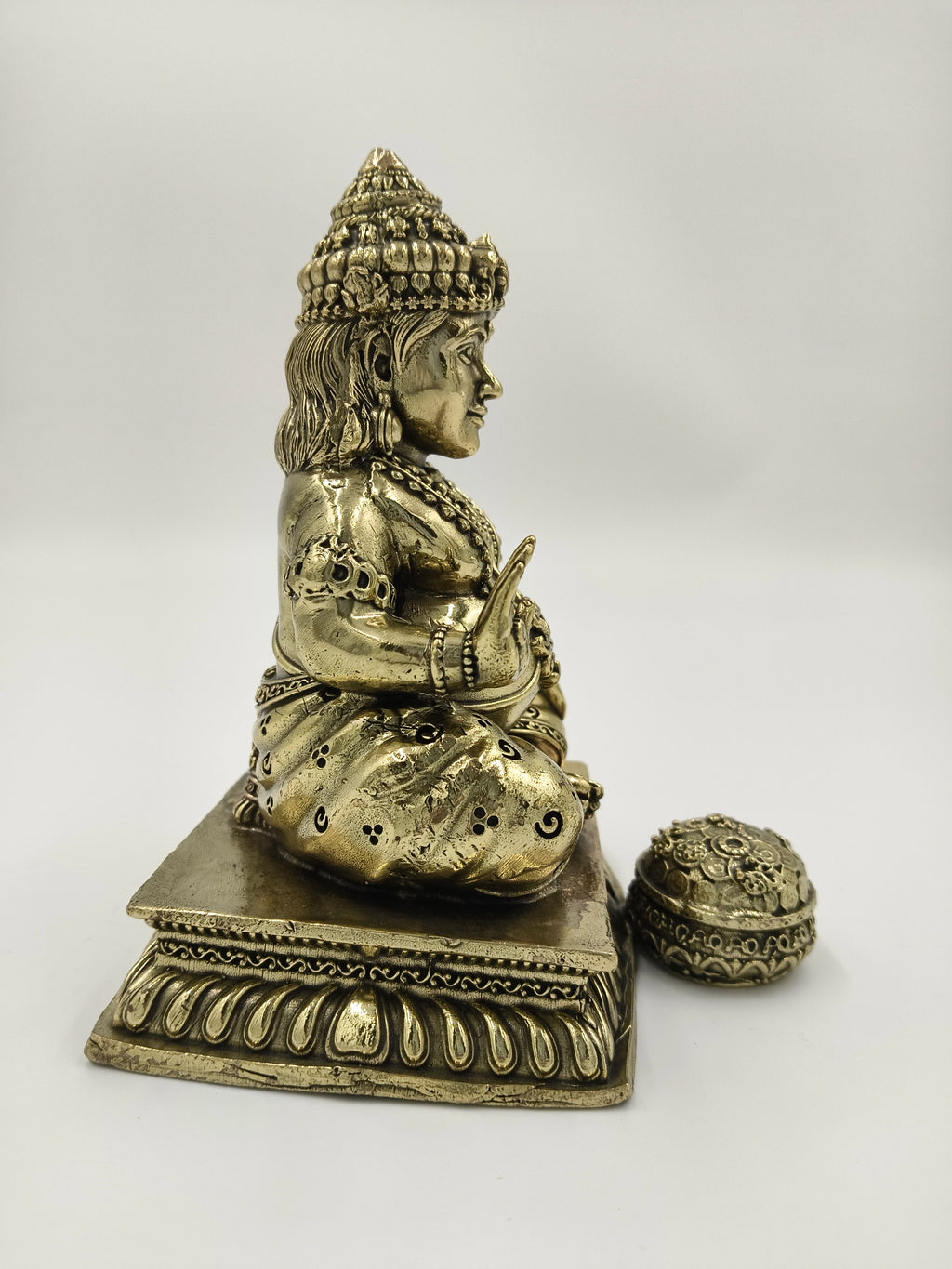 Side view of medium brass Kubera statue seated on decorative pedestal with coin pot
