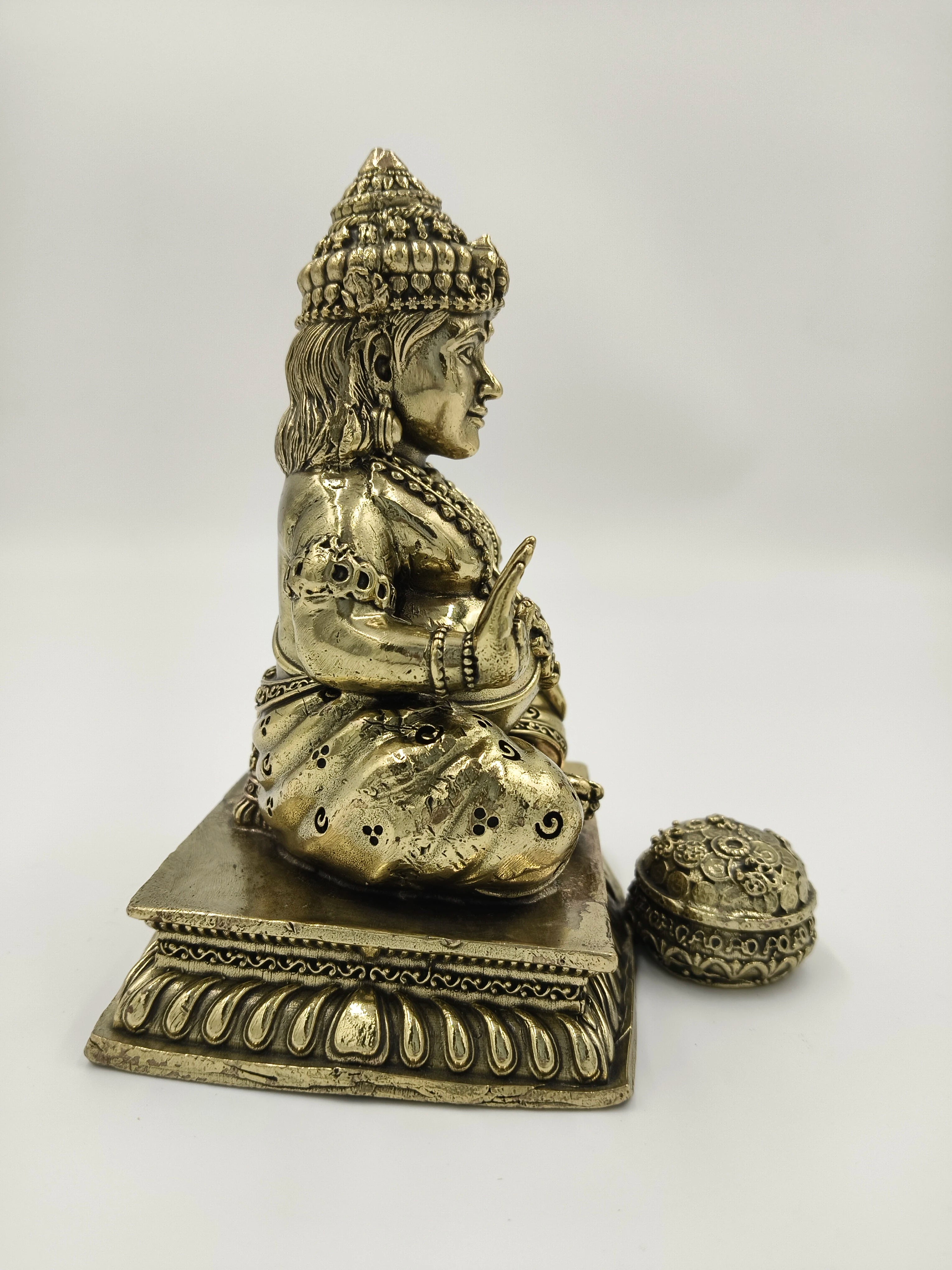 Side view of medium brass Kubera statue seated on decorative pedestal with coin pot