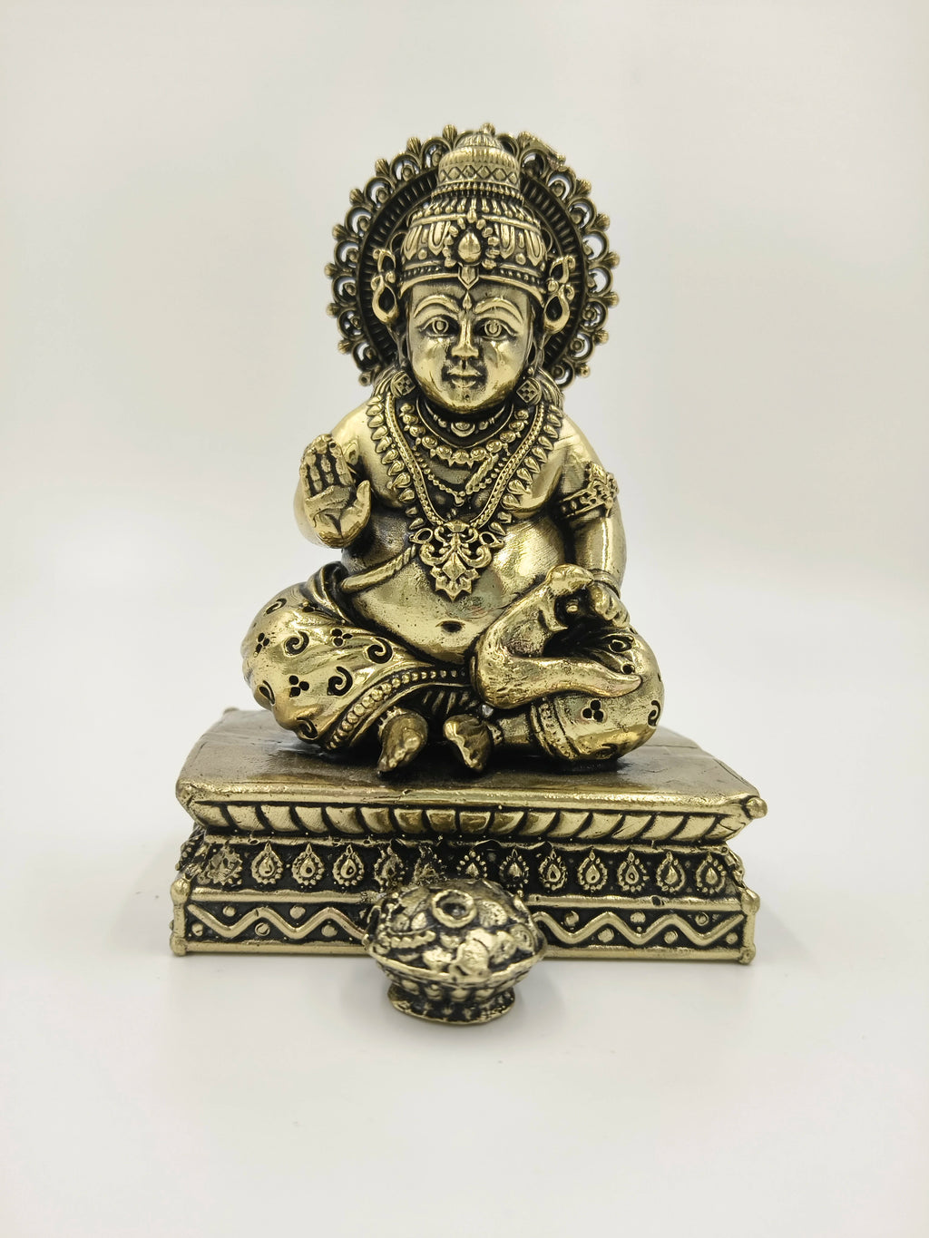 Small brass Kubera sculpture seated on ornate pedestal with pot in front