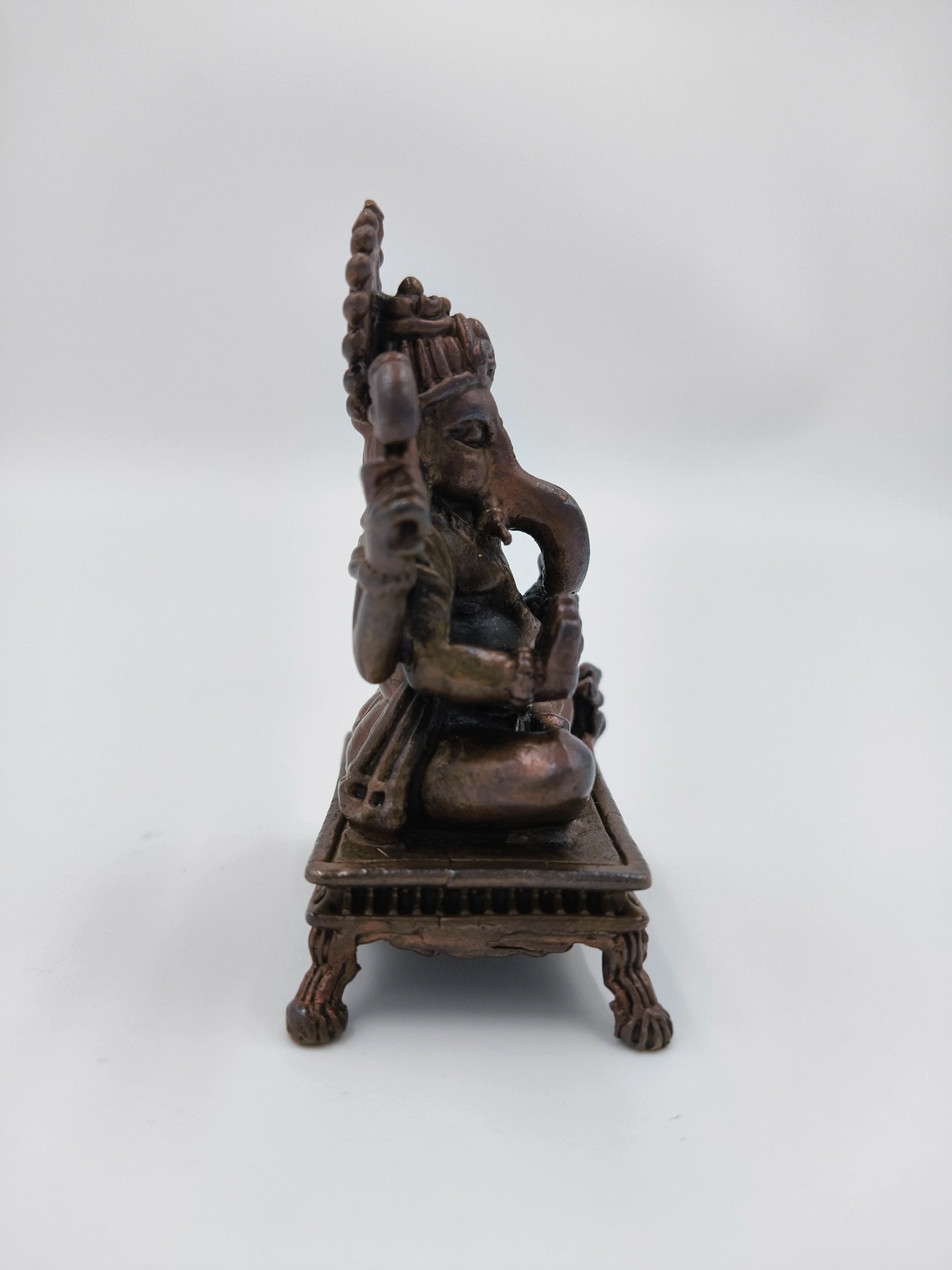 Side view of a small copper Ganesh statue seated on a decorative chowki