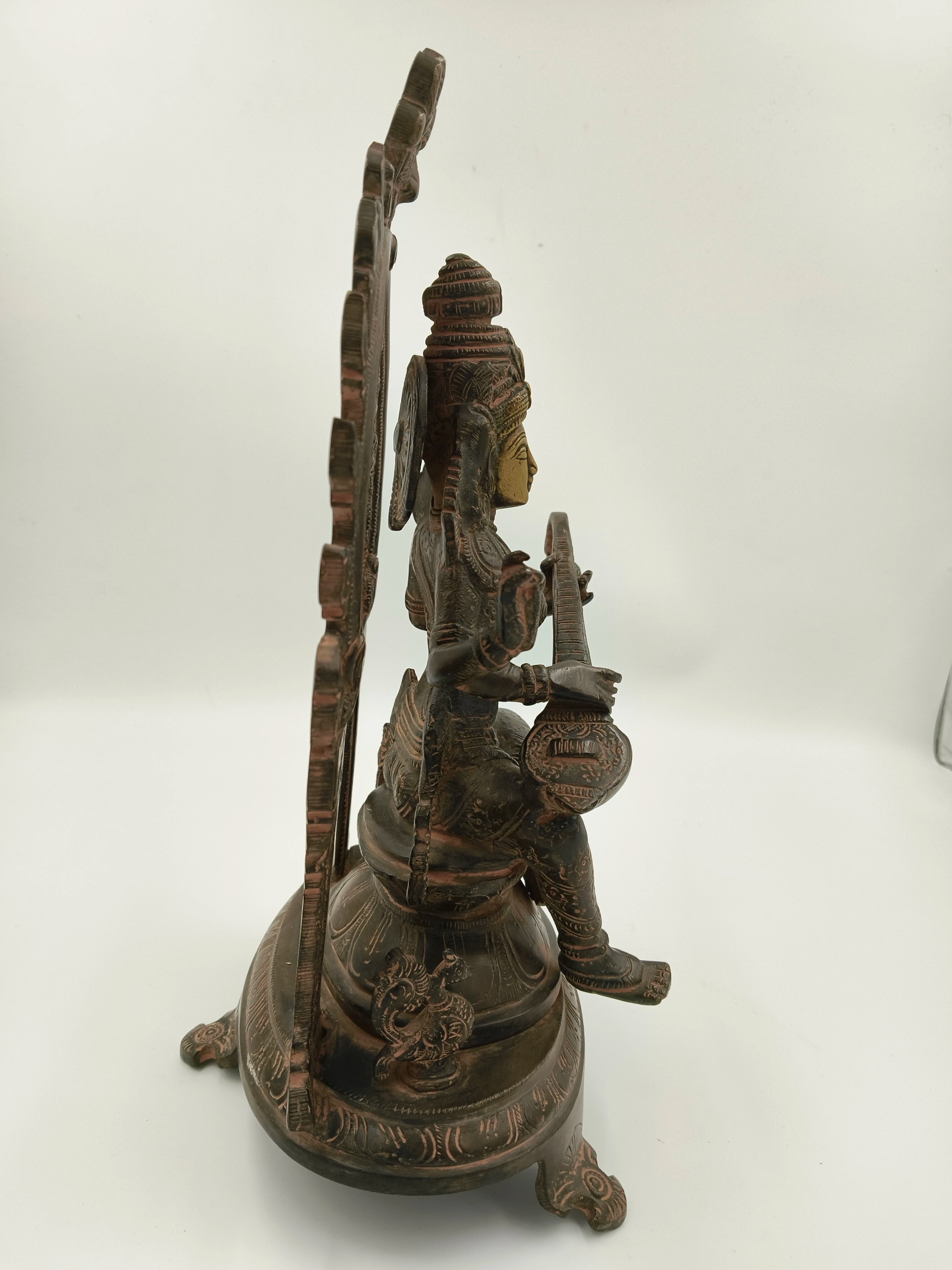 Side view of large brass Saraswathi sculpture seated with arch and musical instrument