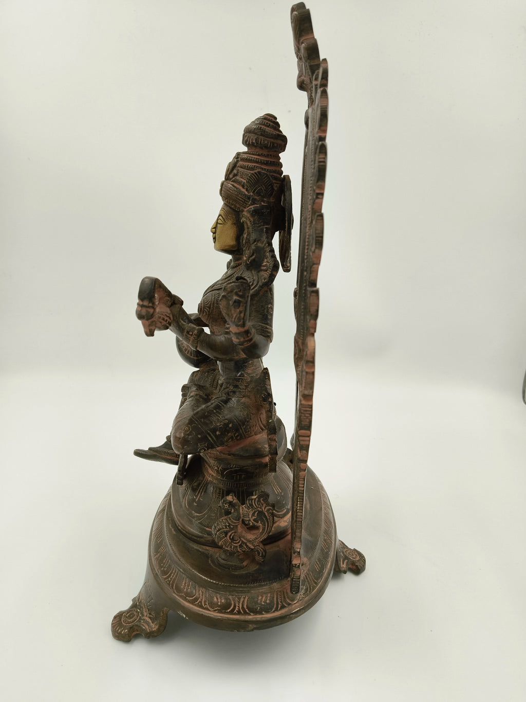 Side view of brass sculpture of Saraswathi sitting with arch and decorative base
