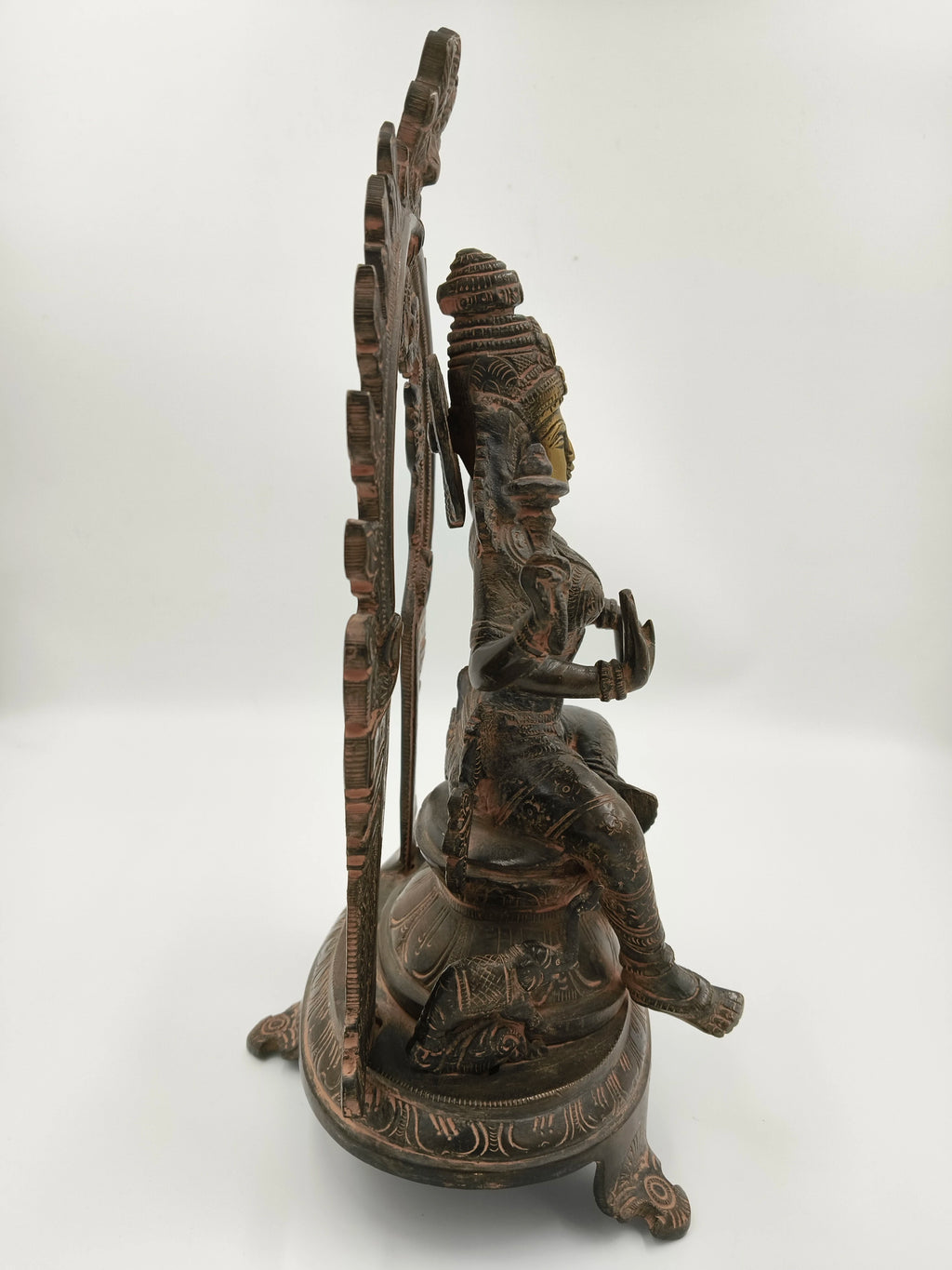 Side view of brass sculpture of deity Lakshmi seated with decorative arch and base