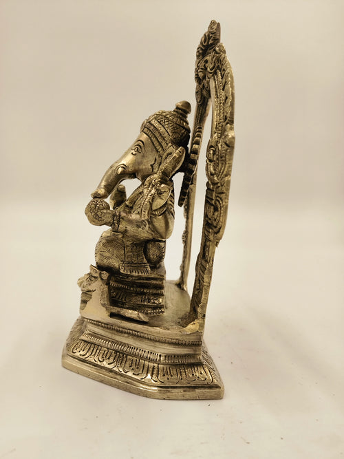 Side view of brass Ganesh sculpture seated on ornate base with decorative arch