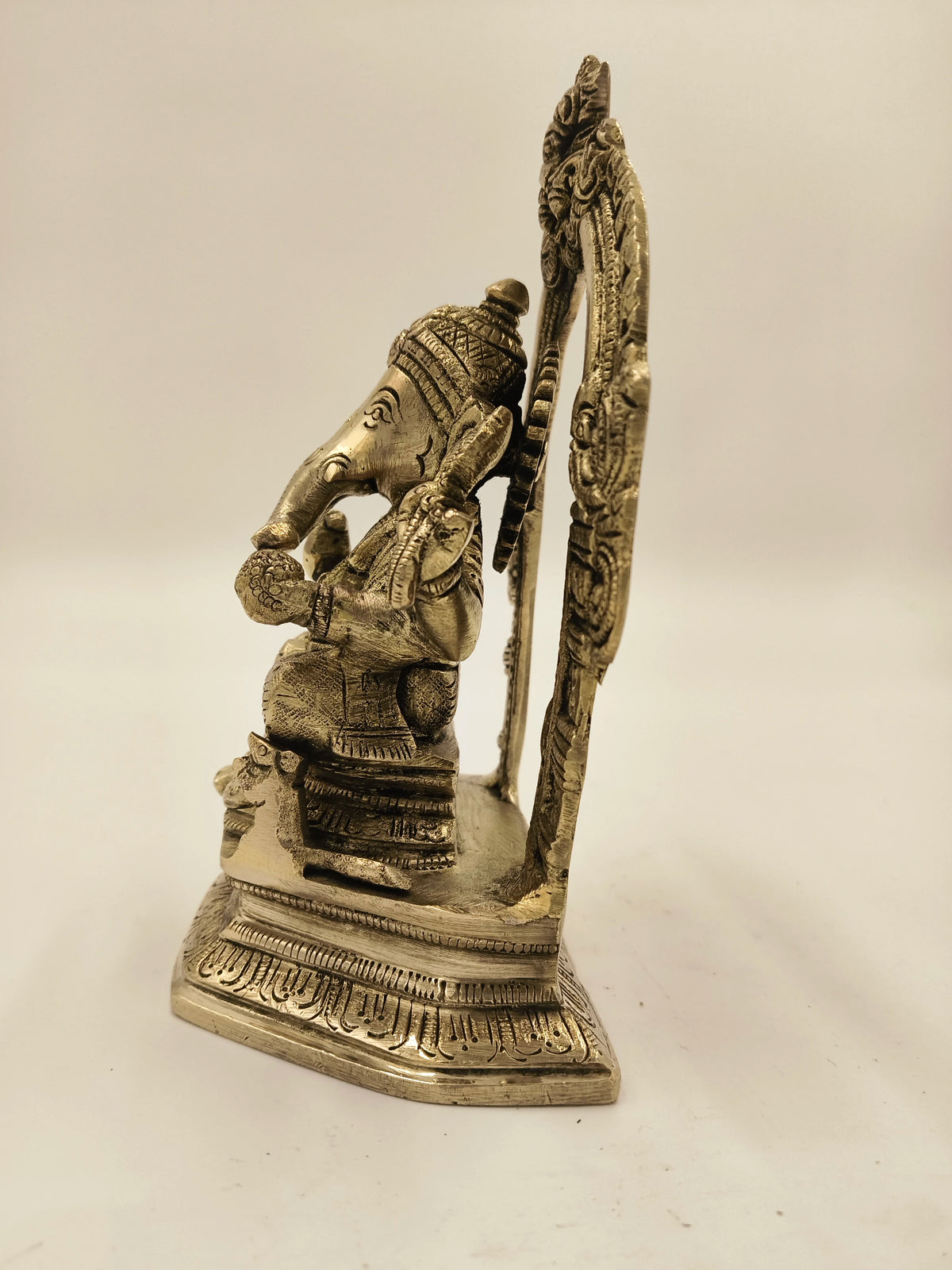 Side view of brass Ganesh sculpture seated on ornate base with decorative arch