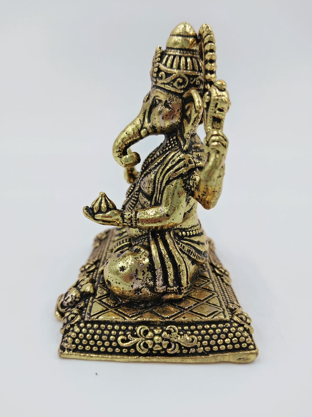 Small brass statue of seated Ganesh holding a sweet on decorative base side view