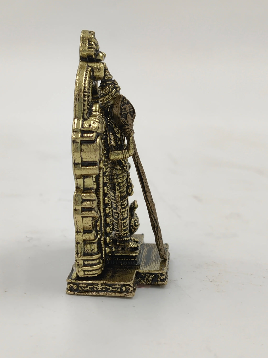 Side view of small brass Murugan statue holding spear with detailed base