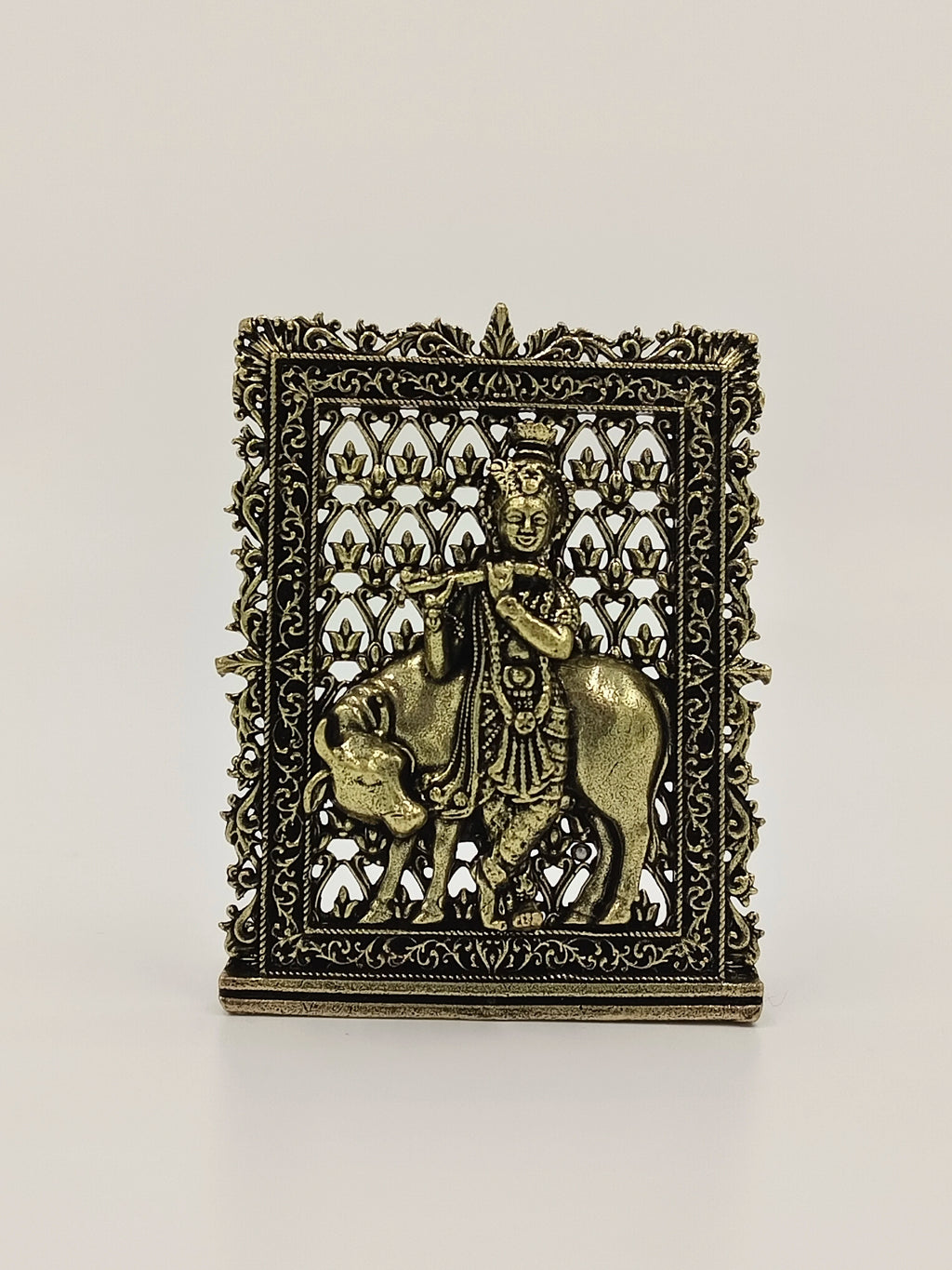 Small brass panel sculpture of Krishna playing flute with cow, intricate floral frame
