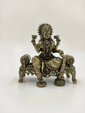 Small brass Lakshmi statue seated on an ornate bench holding lotus flowers