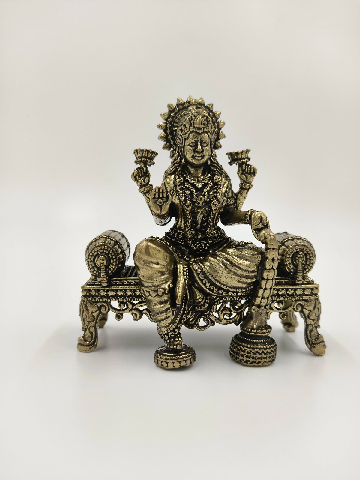 Small brass Lakshmi statue seated on an ornate bench holding lotus flowers