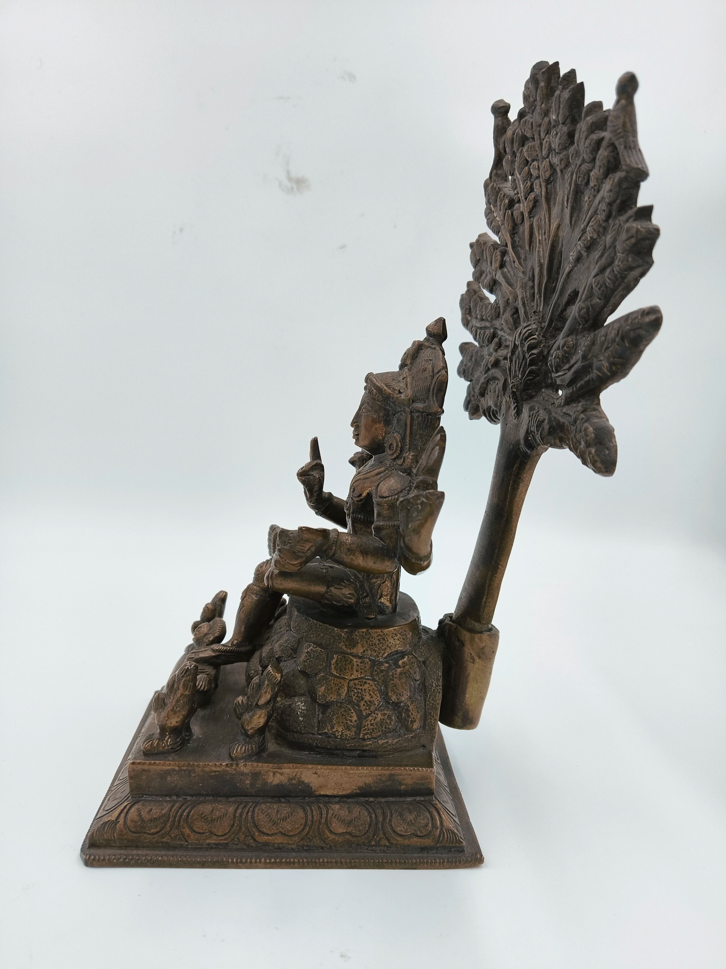 Side view of a bronze Dakshanamurthi sculpture seated with ornate details and a large decorative backdrop
