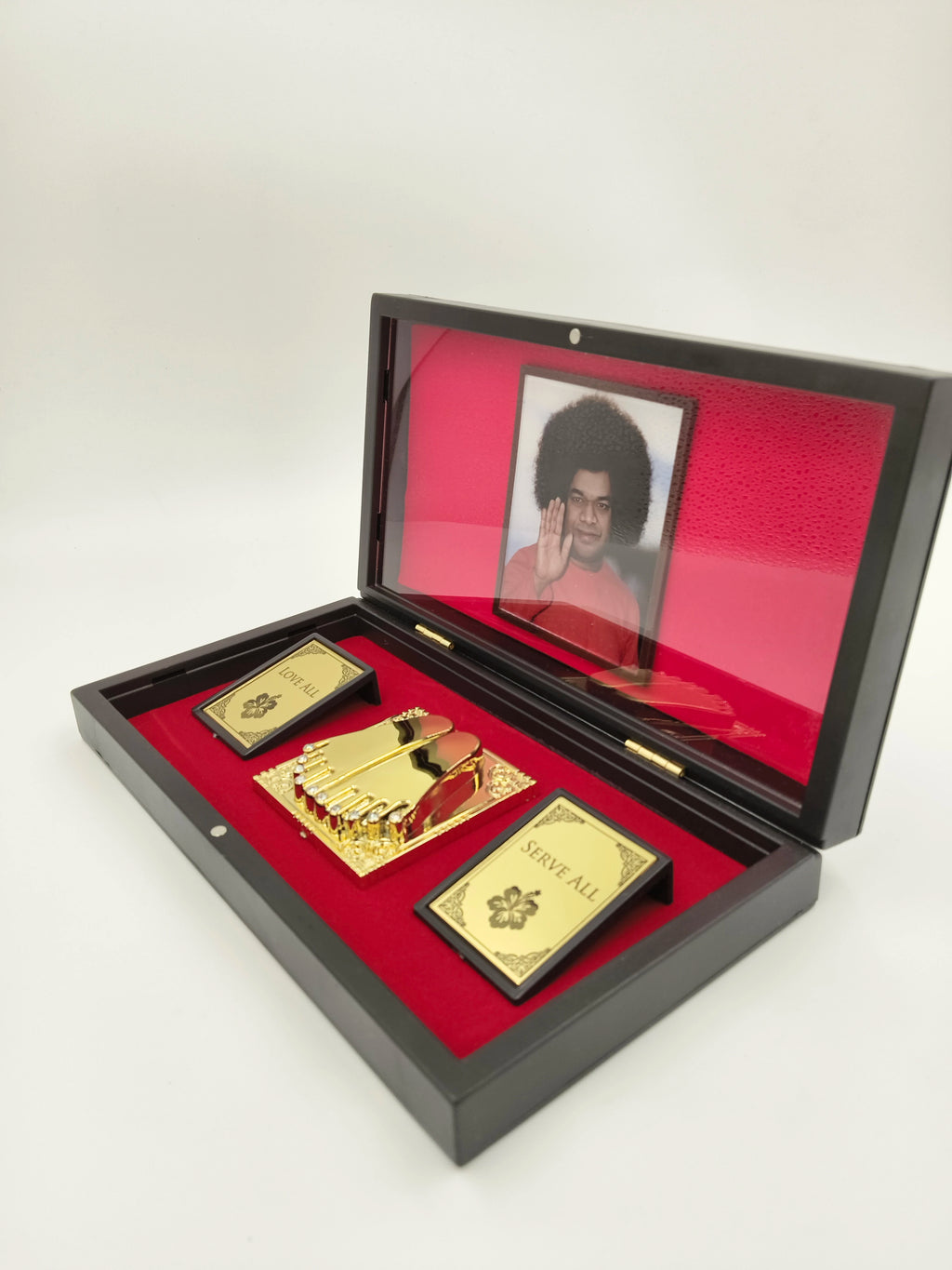 Open black religious box with red interior holding golden Sai Baba paduka and plaques reading Love All and Serve All
