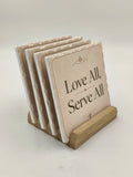 Set of ceramic coasters with 'Love All, Serve All' text on wooden holder